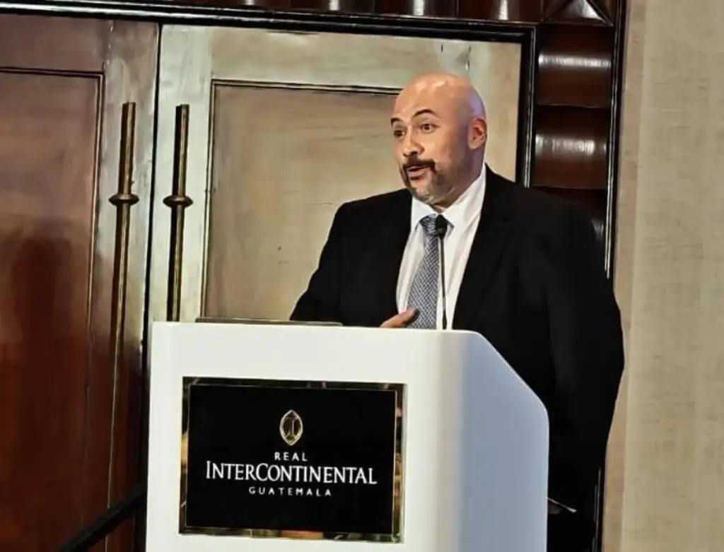 Hombre calvo con traje y corbata hablando en un podio con el logo Real Intercontinental Guatemala, en un evento profesional.