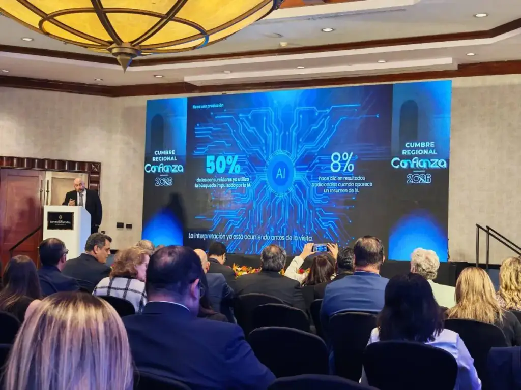 Hombre en podio presenta sobre IA en Cumbre Regional Confianza 2026. Pantalla muestra estadísticas de uso de IA en búsquedas.