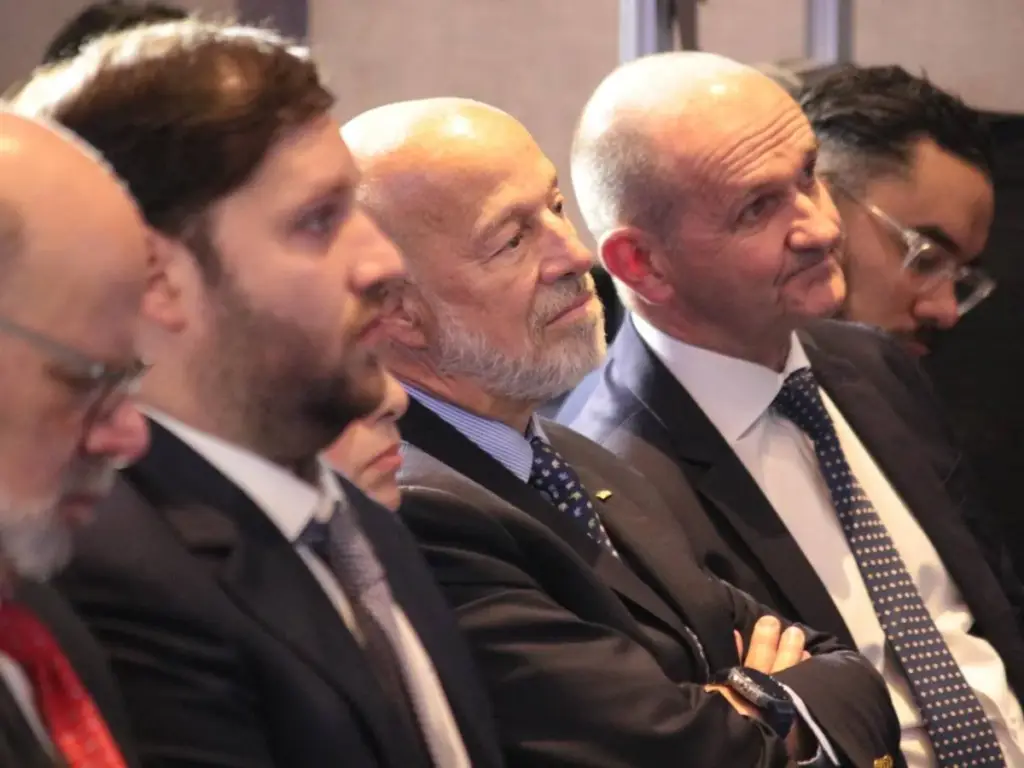 Grupo de hombres profesionales, algunos con barba y gafas, sentados en un evento de negocios, vistiendo trajes y corbatas, prestando atención.