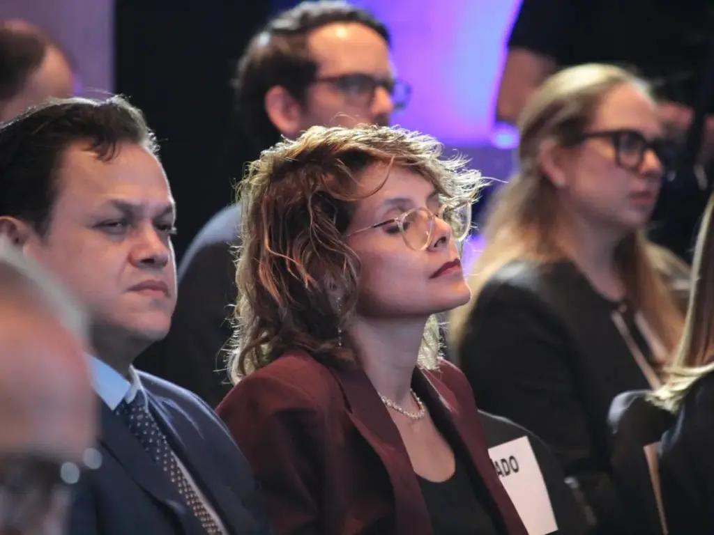 Grupo de profesionales en un evento de negocios. Un hombre con traje y corbata, y una mujer con blazer y gafas, observan atentamente.