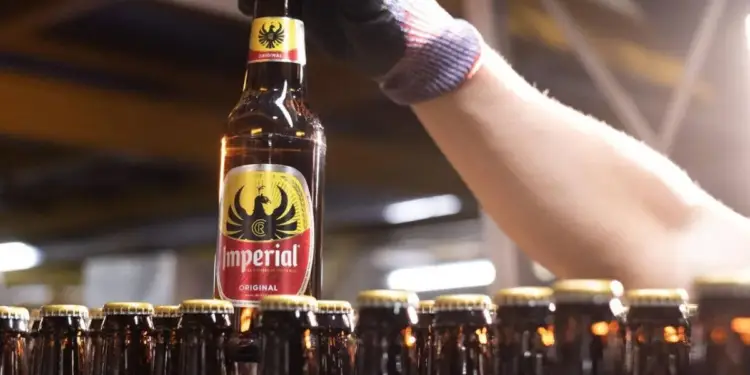 Mano enguantada sosteniendo una botella de cerveza Imperial en una línea de producción. Filas de botellas llenas con tapas doradas.
