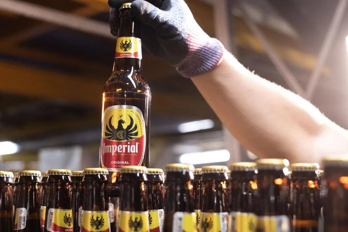 Mano enguantada sosteniendo una botella de cerveza Imperial en una línea de producción. Filas de botellas llenas con tapas doradas.