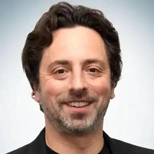 Retrato de un hombre sonriente con cabello oscuro y ondulado, barba corta y ojos claros. Viste una chaqueta oscura y una camisa negra, sobre un fondo degradado claro.
