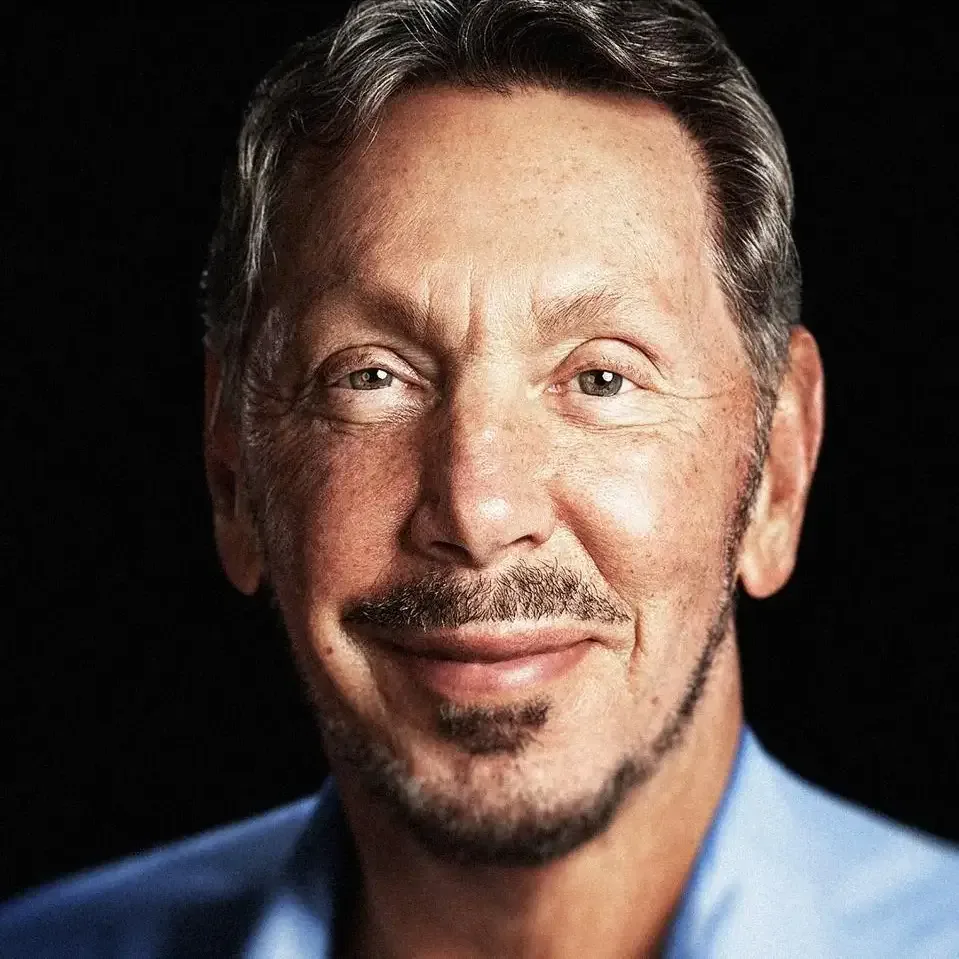 Primer plano de un hombre caucásico de edad avanzada con cabello gris, bigote y barba, vistiendo una camisa azul claro, sonriendo ligeramente con un fondo oscuro. Transmite profesionalismo y experiencia en el ámbito empresarial.