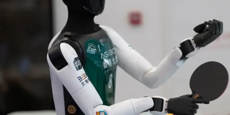 Primer plano de un robot humanoide con un chaleco verde sosteniendo una raqueta de tenis de mesa y una pelota naranja.