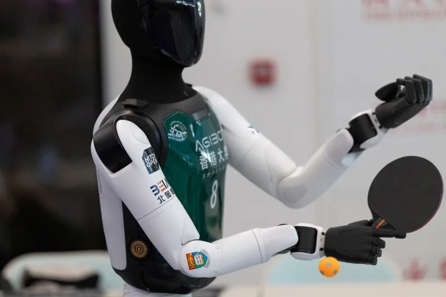Primer plano de un robot humanoide con un chaleco verde sosteniendo una raqueta de tenis de mesa y una pelota naranja.