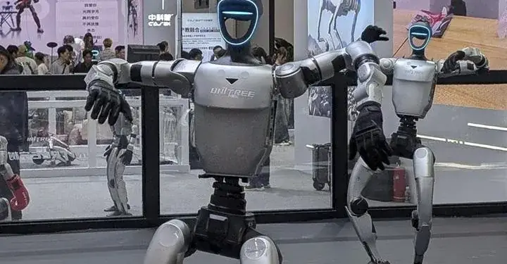 Dos robots humanoides de pie y con luces azules en la cabeza en una feria tecnológica, mostrando avances de la robótica.