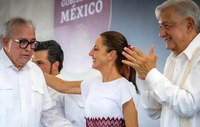 Tres personas en un evento oficial. Un hombre mayor sonríe mientras gesticula cerca de una mujer que ríe. Otro hombre con gafas observa. Fondo con 'Gobierno México'.
