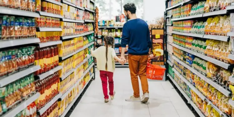 Un hombre y una niña caminan de la mano por un pasillo de supermercado lleno de estantes con productos. El hombre lleva una cesta roja.