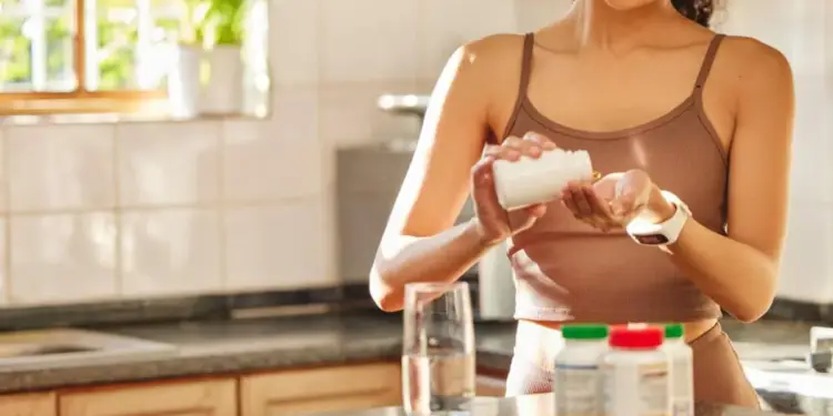 Mujer vertiendo suplementos de un frasco blanco en su mano, con un vaso de agua y otros frascos en una encimera de cocina.