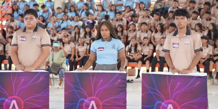Tres estudiantes de pie frente a atriles con gráficos de plasma, mientras una audiencia de jóvenes los observa sentados en gradas. Un proyector está visible en primer plano.