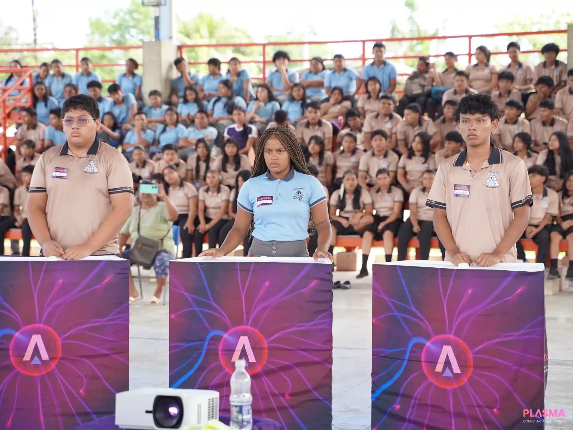 Tres estudiantes de pie frente a atriles con gráficos de plasma, mientras una audiencia de jóvenes los observa sentados en gradas. Un proyector está visible en primer plano.