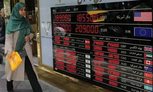 Una mujer con velo verde y abrigo beige mira un tablero electrónico que muestra tasas de compra y venta de divisas extranjeras como USD, EUR, GBP, AED, CAD, AUD, TRY, CHF, SEK y NOK.