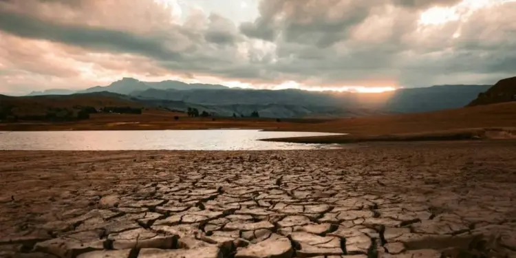 Vista de un paisaje árido con tierra agrietada en primer plano, un cuerpo de agua reducido y montañas al fondo bajo un cielo nublado con el sol poniente.