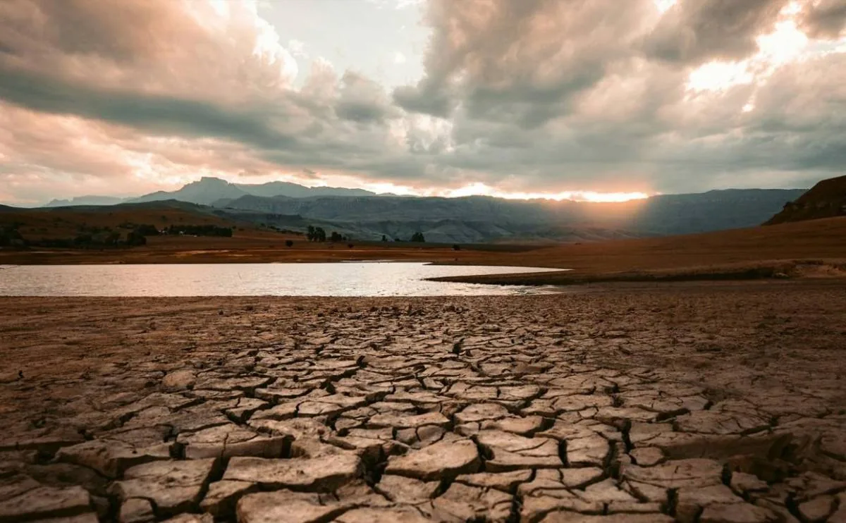 Vista de un paisaje árido con tierra agrietada en primer plano, un cuerpo de agua reducido y montañas al fondo bajo un cielo nublado con el sol poniente.