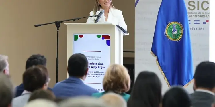 Una mujer con traje blanco habla desde un podio frente a una audiencia atenta y una bandera azul.