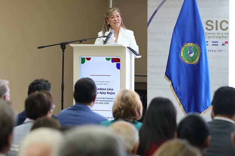 Una mujer con traje blanco habla desde un podio frente a una audiencia atenta y una bandera azul.