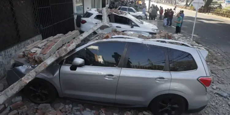 Dos vehículos, un SUV plateado y uno blanco, aplastados por escombros de ladrillo y concreto de un edificio colapsado en una calle urbana. Personas observan la escena.