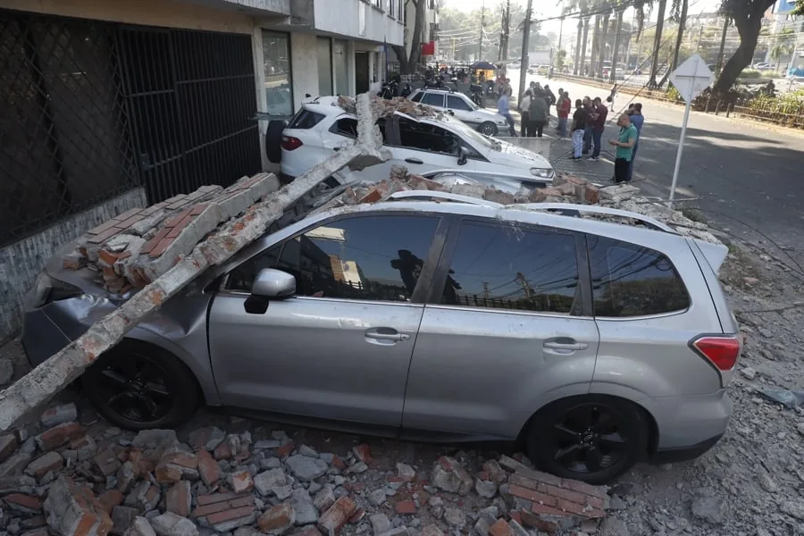 Dos vehículos, un SUV plateado y uno blanco, aplastados por escombros de ladrillo y concreto de un edificio colapsado en una calle urbana. Personas observan la escena.