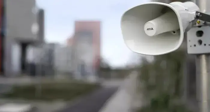 Primer plano de un altavoz o bocina de seguridad de color blanco montado en un poste metálico con edificios difuminados al fondo.