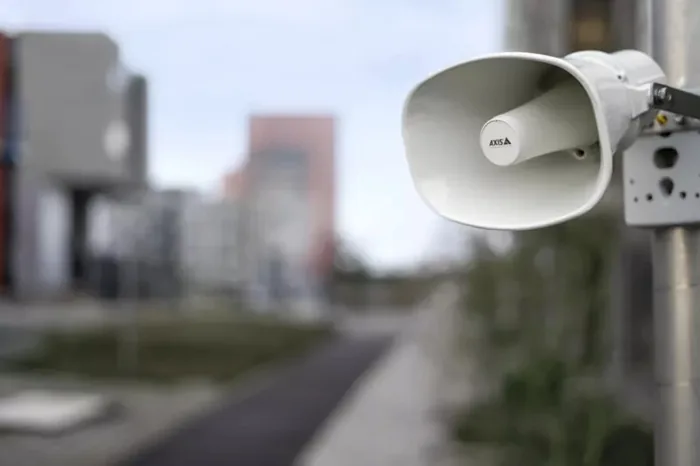 Primer plano de un altavoz o bocina de seguridad de color blanco montado en un poste metálico con edificios difuminados al fondo.
