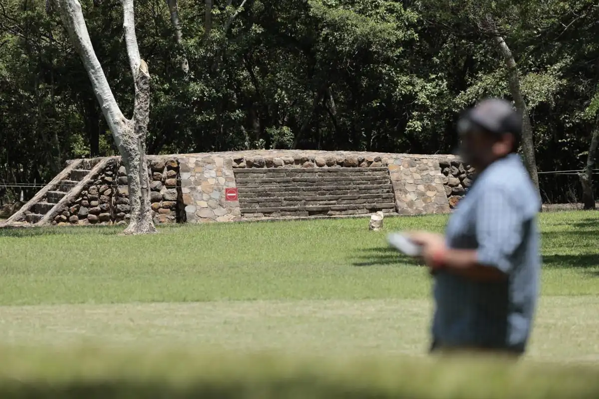 Estructura de piedra antigua con escalones y un visitante desenfocado sosteniendo un folleto en un área verde con árboles.