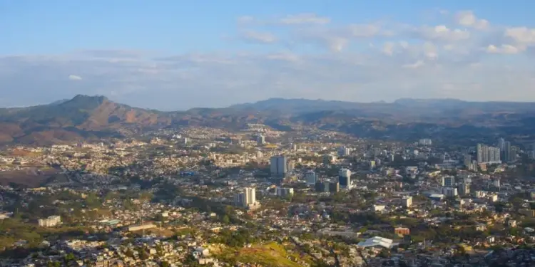 Vista aérea panorámica de una extensa ciudad en un valle, rodeada por montañas y colinas, con edificios residenciales y comerciales bajo un cielo azul claro.