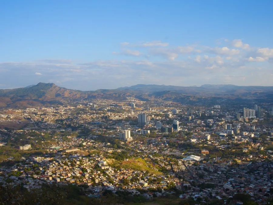 Vista aérea panorámica de una extensa ciudad en un valle, rodeada por montañas y colinas, con edificios residenciales y comerciales bajo un cielo azul claro.