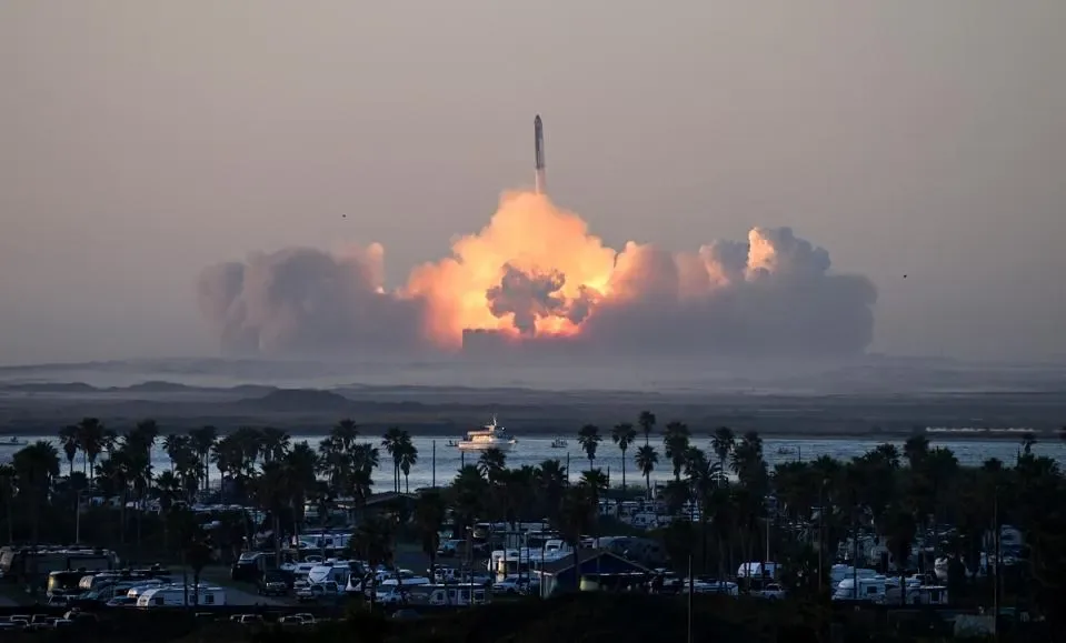 Un cohete espacial despega generando una enorme nube de fuego y humo al fondo, visto desde una zona costera con palmeras.