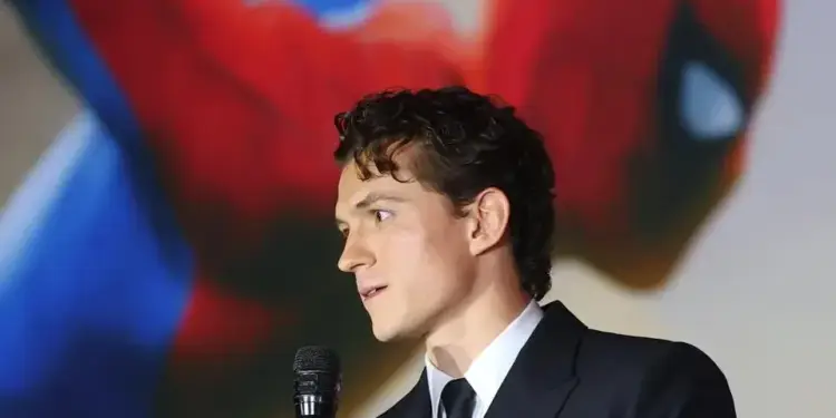 Hombre joven de perfil en traje oscuro y corbata, hablando en un micrófono. Fondo borroso rojo y azul con figura.