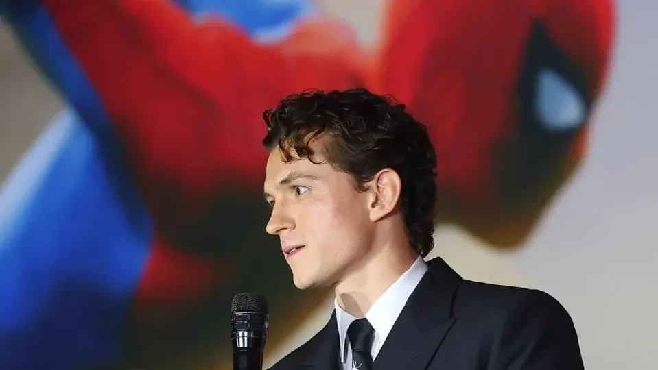 Hombre joven de perfil en traje oscuro y corbata, hablando en un micrófono. Fondo borroso rojo y azul con figura.