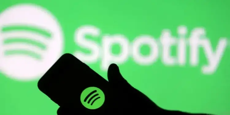Silueta de una mano sosteniendo un smartphone que muestra el logotipo de Spotify frente a una pantalla verde con el nombre de la marca.