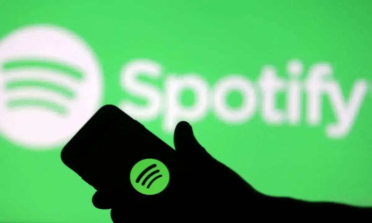Silueta de una mano sosteniendo un smartphone que muestra el logotipo de Spotify frente a una pantalla verde con el nombre de la marca.