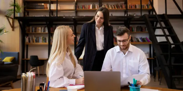 Tres profesionales reunidos alrededor de una mesa de madera trabajando con una laptop y documentos en una oficina con estanterías llenas de libros al fondo.