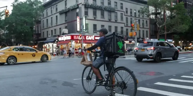 Repartidor en bicicleta con mochila de entrega y bolsa de papel en un cruce urbano. Edificios, un taxi amarillo y un SUV gris de fondo.