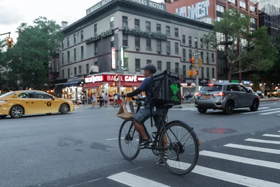Repartidor en bicicleta con mochila de entrega y bolsa de papel en un cruce urbano. Edificios, un taxi amarillo y un SUV gris de fondo.