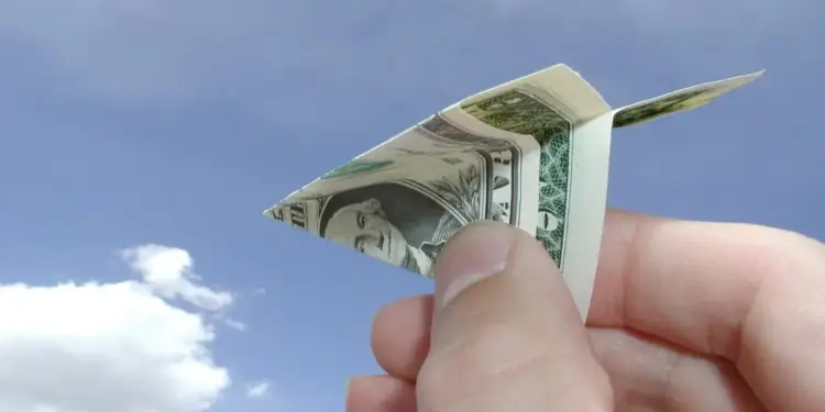Mano sosteniendo un avión de papel hecho con un billete de dólar estadounidense contra un cielo azul con nubes.