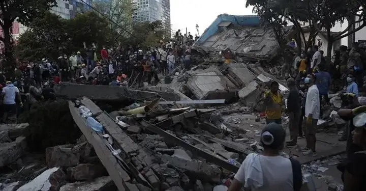 Escena de desastre urbano con un edificio colapsado, grandes pilas de escombros y numerosas personas en labores de rescate y apoyo. Edificios modernos al fondo.