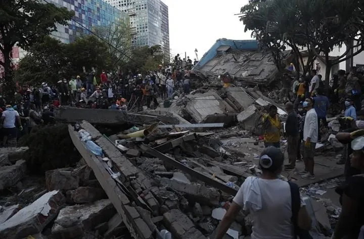 Escena de desastre urbano con un edificio colapsado, grandes pilas de escombros y numerosas personas en labores de rescate y apoyo. Edificios modernos al fondo.