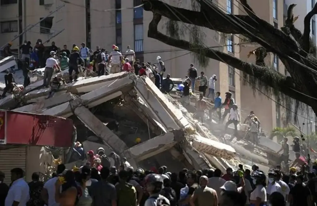 Vista de un edificio colapsado con escombros de hormigón y acero. Numerosas personas, incluyendo rescatistas, trabajan entre los restos. Polvo en el aire. Un gran árbol a la derecha.