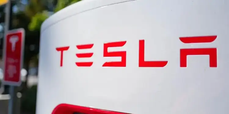 Primer plano del logotipo rojo 'TESLA' en una estación de carga blanca para vehículos eléctricos. Al fondo, un poste con un cartel rojo borroso.