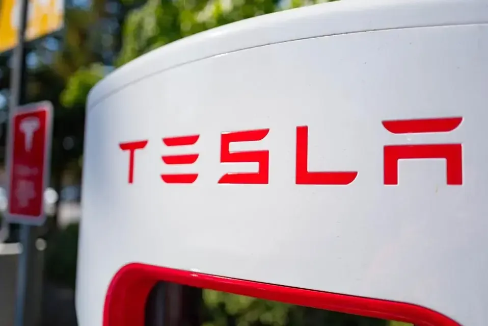 Primer plano del logotipo rojo 'TESLA' en una estación de carga blanca para vehículos eléctricos. Al fondo, un poste con un cartel rojo borroso.