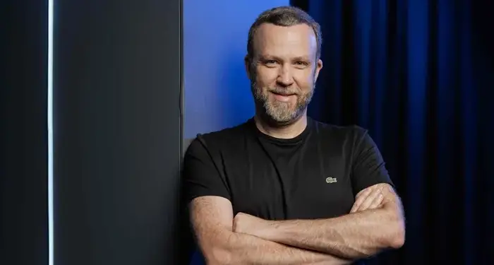 Hombre profesional con barba y camiseta negra, brazos cruzados, sonriendo. Fondo azul oscuro y pared gris con luz.