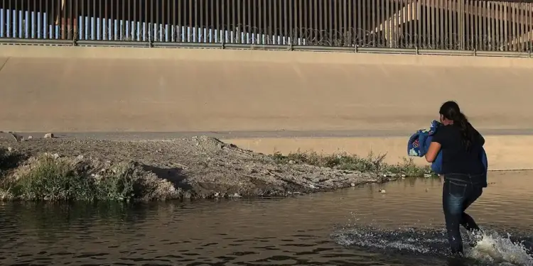 Mujer cruzando un río con un bulto en brazos, bajo una valla fronteriza con alambre de púas.