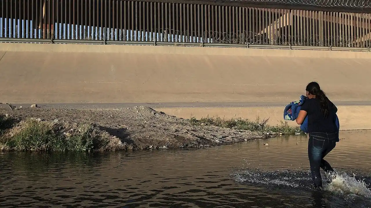 Mujer cruzando un río con un bulto en brazos, bajo una valla fronteriza con alambre de púas.