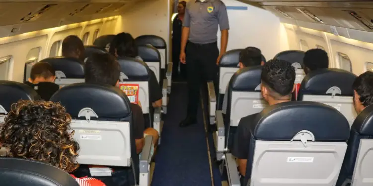 Pasajeros sentados en el interior de un avión comercial mientras un oficial en uniforme vigila el pasillo.