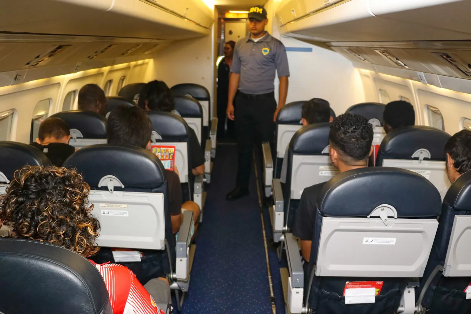 Pasajeros sentados en el interior de un avión comercial mientras un oficial en uniforme vigila el pasillo.
