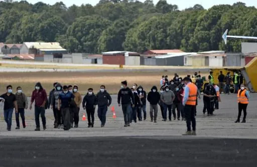 Grupo de personas con mascarillas caminando en una pista de aeropuerto bajo supervisión. Personal con chalecos naranjas y edificios al fondo.