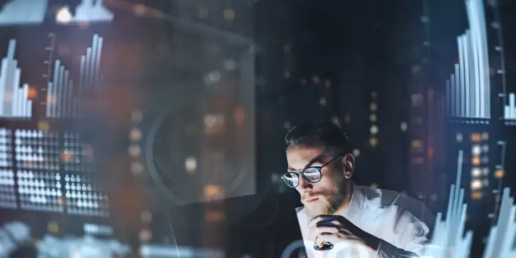 Hombre con gafas trabajando de noche rodeado de gráficos estadísticos y datos financieros proyectados en pantallas.
