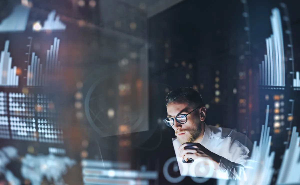 Hombre con gafas trabajando de noche rodeado de gráficos estadísticos y datos financieros proyectados en pantallas.